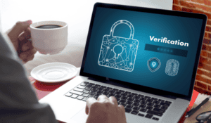 A laptop sitting on a desk with a padlock displayed on the screen. A man's right hand is seen hovering above the keyword, his left hand is holding a cup of coffee.