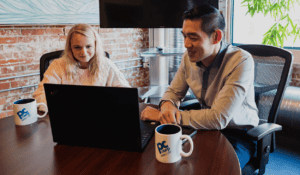 Two IT professionals, a man and a woman, seated at a desk discussing cybersecurity strategies, with PC Corp branded mugs and computers in an office with brick walls.