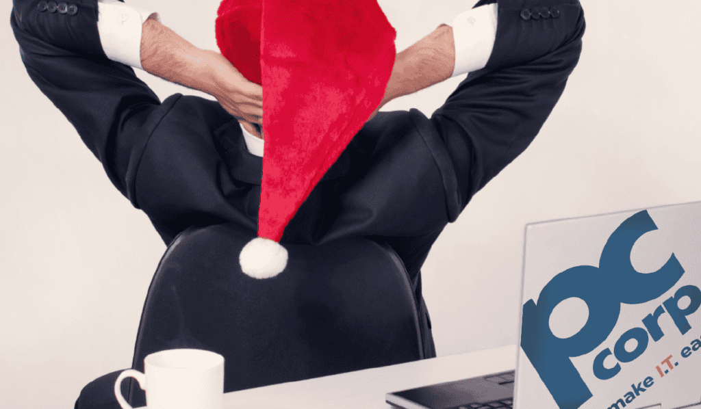 A professional in a suit sitting backwards on a chair, whimsically wearing a Santa hat, in an office setting, with a laptop with PC Corp's logo on it