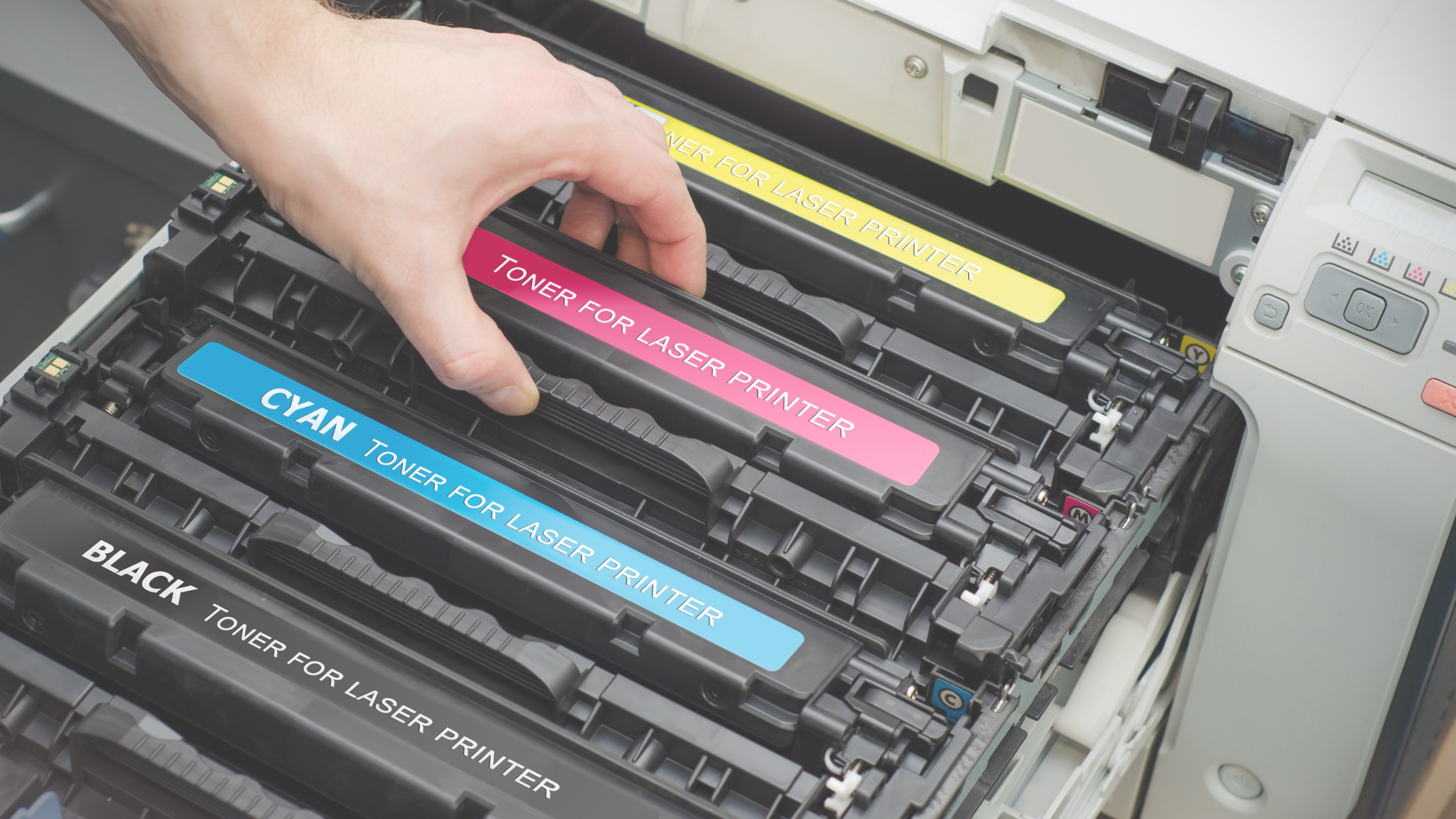 Close-up of a person's hand replacing toner cartridges in a laser printer. There are four cartridges visible labeled with their respective colors: black, cyan, magenta, and yellow. The printer is open, showing the internal cartridge housing, indicating routine maintenance or replacement of printing supplies.