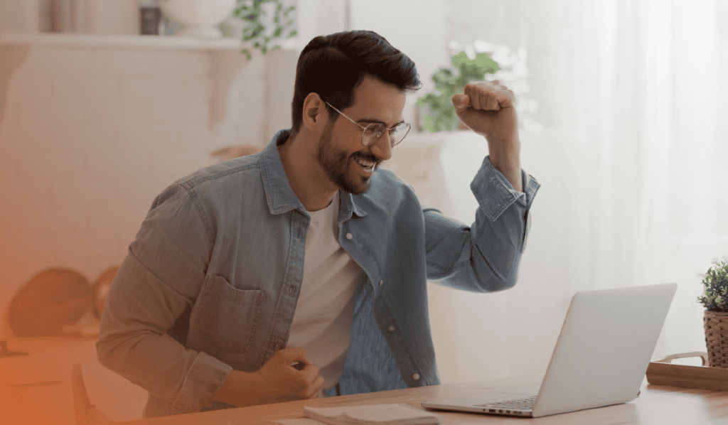 A joyful man in glasses, sitting at a home office desk, celebrating success while looking at his laptop screen. His expression is one of triumph, raising his fist in the air in a cheerful victory gesture. The setting is light and modern, with a comfortable home office vibe.