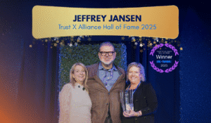 A photo representing the theme/subject of the blog "A Lifetime of IT Leadership and Collaboration - PC Corp's Jeffrey Jansen Inducted into the TXA Hall of Fame." There is a photo of Jeffrey standing with two women at the award ceremony, They are all smiling at the camera. They are surrounded by blue curtains, a green backdrop, and the top of the image has Jeffrey's name, award title and a collection of blue/gold stars dotting the background.