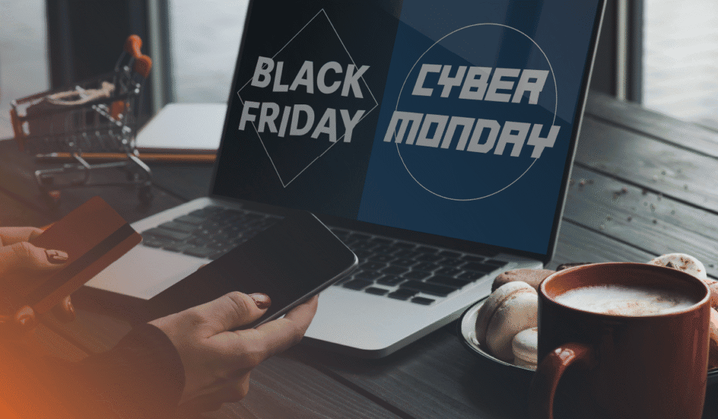 An image representing the title of the blog "Beat Cybersecurity Scams This Black Friday, Cyber Monday and Beyond" with a dark blue, grey and orange theme. Some is holding multiple devices, also a nod to the main subject of the blog, IT procurement, and their computer screen is split with the text "Black Friday" and "Cyber Monday". There is also a cozy coffee cup to the side and a plate of cookies, as well as a mini-shopping cart on the desk.