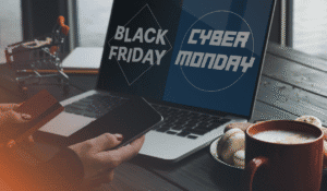 An image representing the title of the blog "Beat Cybersecurity Scams This Black Friday, Cyber Monday and Beyond" with a dark blue, grey and orange theme. Some is holding multiple devices, also a nod to the main subject of the blog, IT procurement, and their computer screen is split with the text "Black Friday" and "Cyber Monday". There is also a cozy coffee cup to the side and a plate of cookies, as well as a mini-shopping cart on the desk.