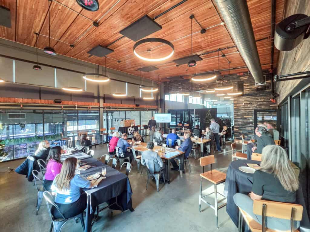 Attendees sit at various tables in a crowded room at the brewery, all facing the front where there is a presentation screen and Christmas tree.