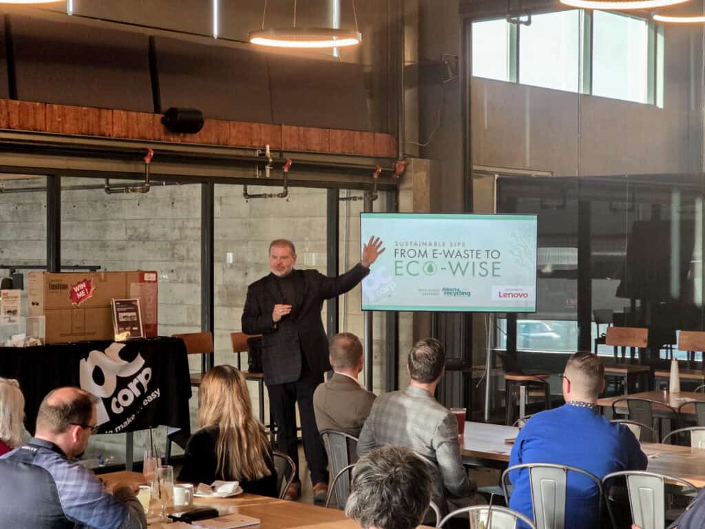 A man wearing all black waves at the audience while speaking, in front of a screen with the event title, Sustainability Sips: From E-Waste to Eco-Wise, and next to a table with the PC Corp logo and giveaway items.