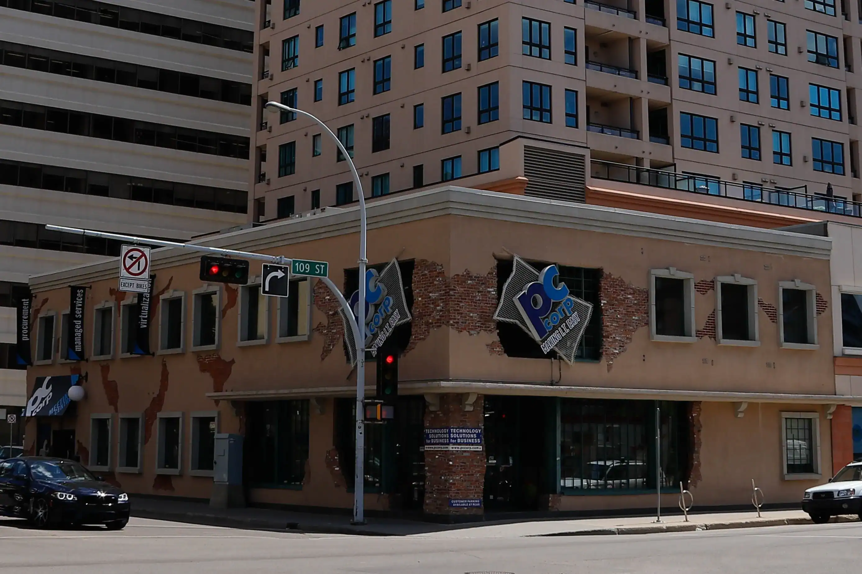 An image of the PC Corp office building, it is a short brown building in front of larger office towers, the blue PC Corp logo is large at the corner.