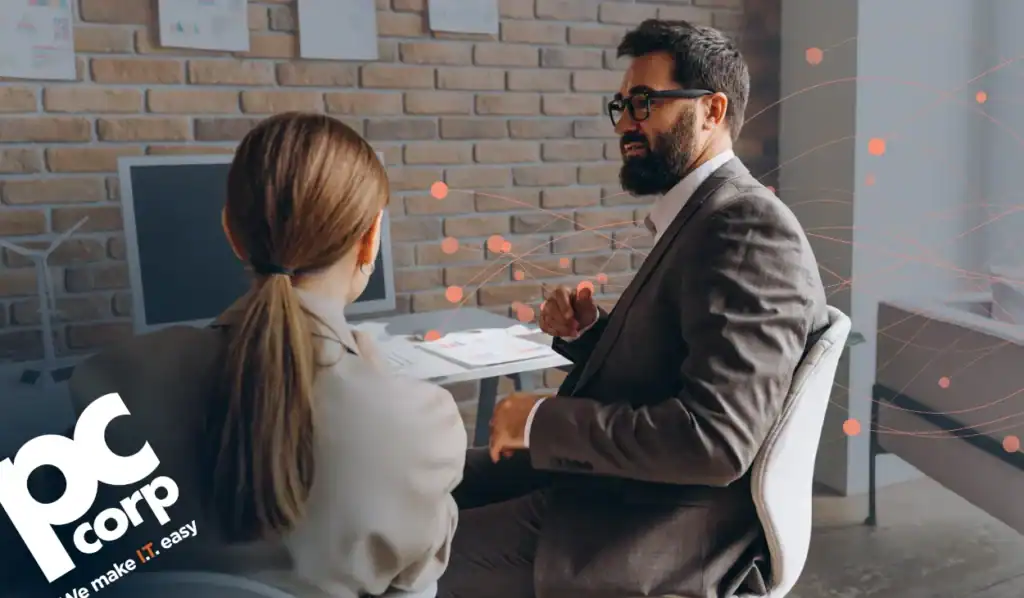 An image to represent the blog title: Local IT Partner vs. National Reseller: What You're Actually Giving Up . A man and a woman in business attire sit in an office, facing away from the camera towards a desk with a computer against a brick wall.