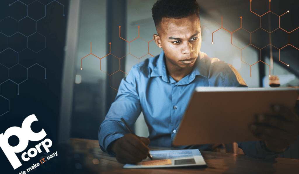 A professional in a blue shirt studies a tablet while taking notes on a printed document at his desk, representing an IT partner carefully evaluating technology vendors.