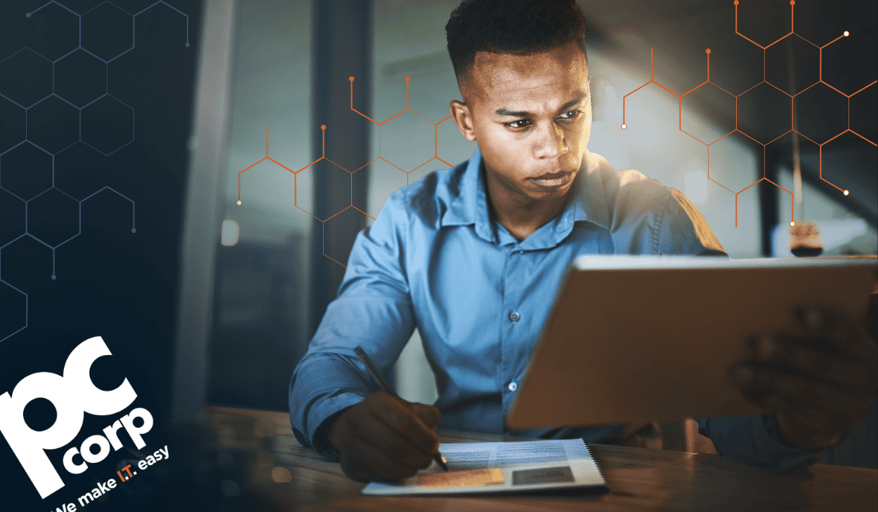 A professional in a blue shirt studies a tablet while taking notes on a printed document at his desk, representing an IT partner carefully evaluating technology vendors.